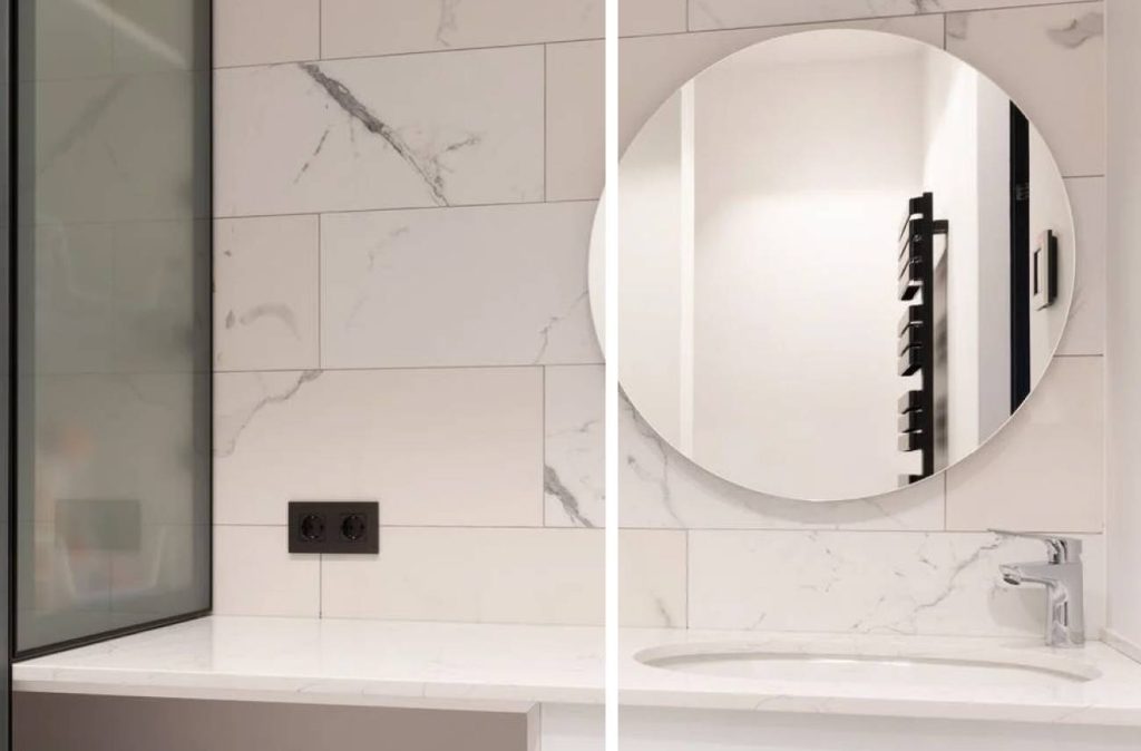 a rounded frameless mirror with tiled marble backsplash and a white bathroom countertop with sink