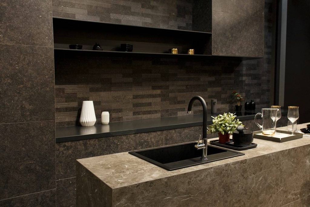 Dark-themed kitchen that features a gray kitchen island with black sink, black faucet, decorative plant, mortar and pestle, and tray with glasses and pitcher on it
