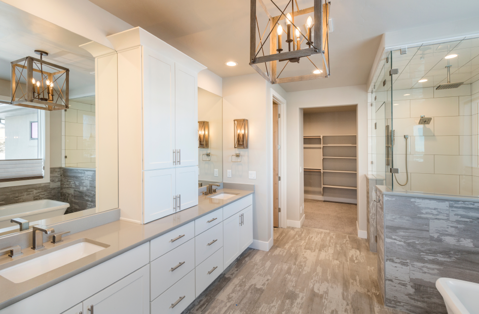 a spacious bathroom with gray flooring and a shower area acroos the vanity area which has two mirrors and sinks separated by a white shelf space that sits on top of the beige bathroom countertop