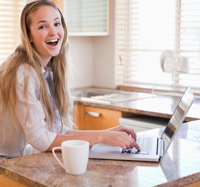 a woman working in a kitchen, perched over the countertop with a laptop and coffee mug